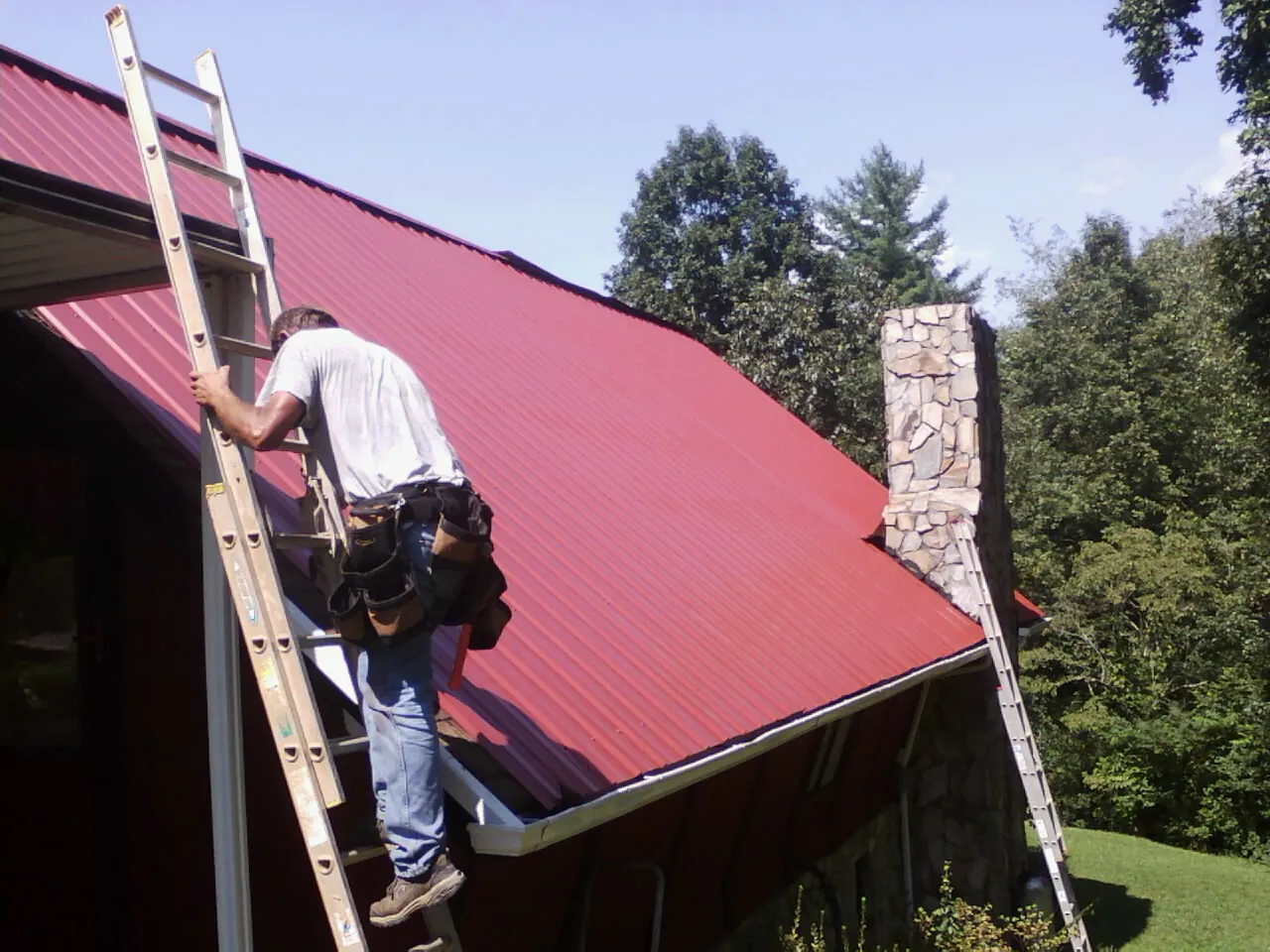 Roofer on a ladder beside a red metal roof and stone chimney — Soffit & Fascia Repair by Roofing Brothers in Broadbent